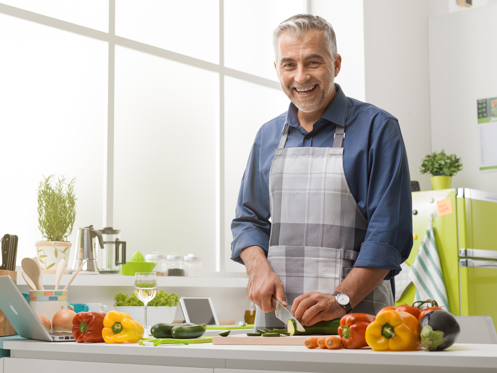 Mature man cooking in the kitchen at home, he is wearing an apron and slicing vegetables, healthy eating lifestyle concept